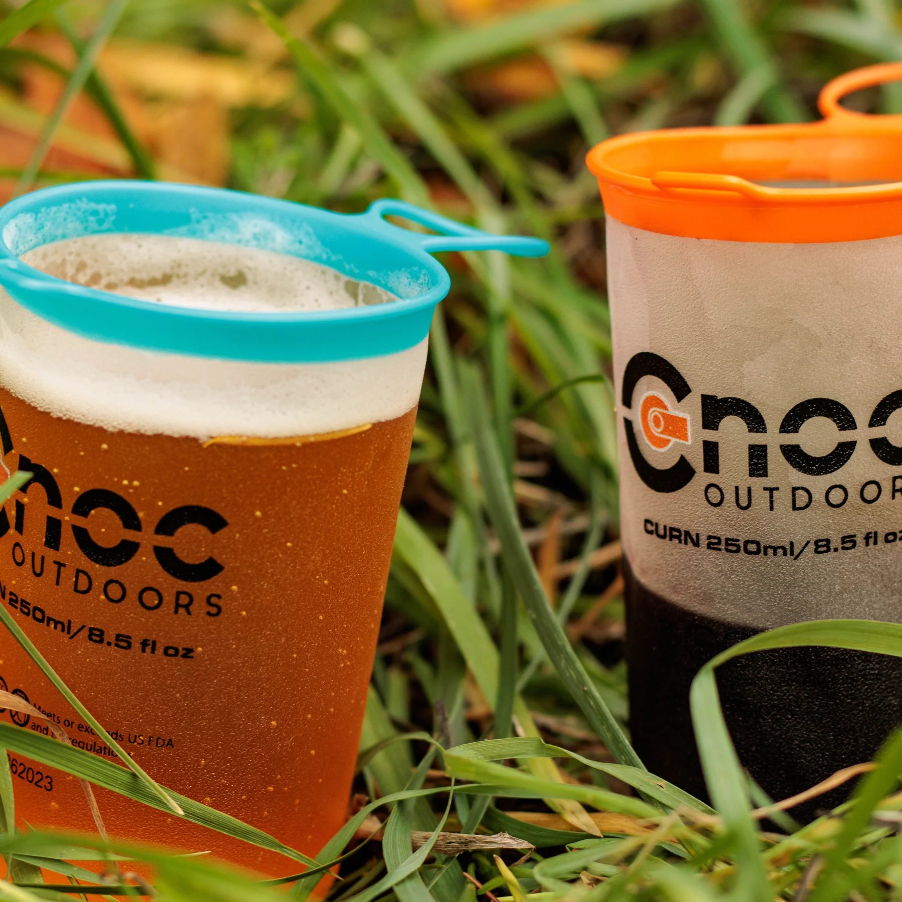Two Cnoc Outdoors CURN 250ml collapsible cups sitting in green grass, one filled with amber beer topped with foam and fitted with a blue silicone rim, the other half filled with wine with an orange rim.