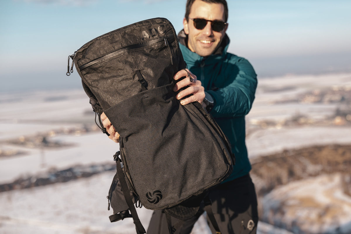 Smiling man holding up a black All Day Carry travel backpack on a wintry hilltop
