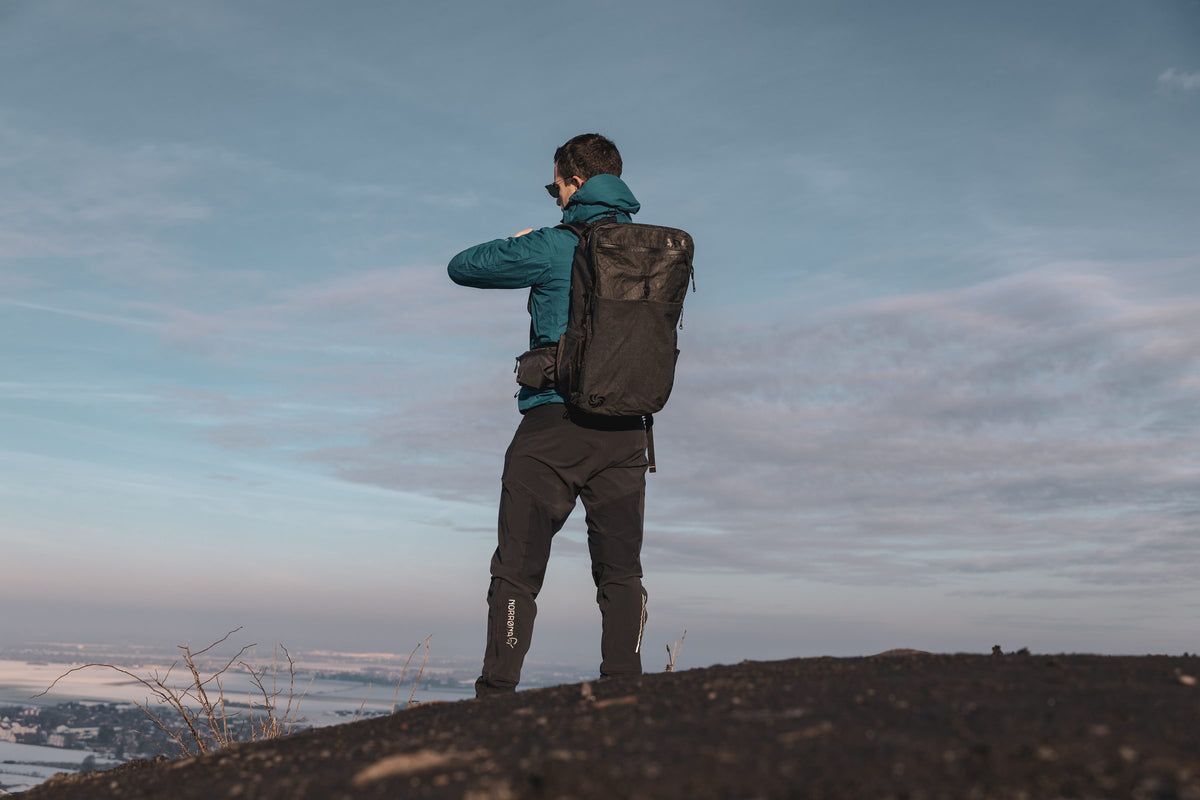 Man wearing a black All Day Carry travel backpack on a hilltop against an evening sky