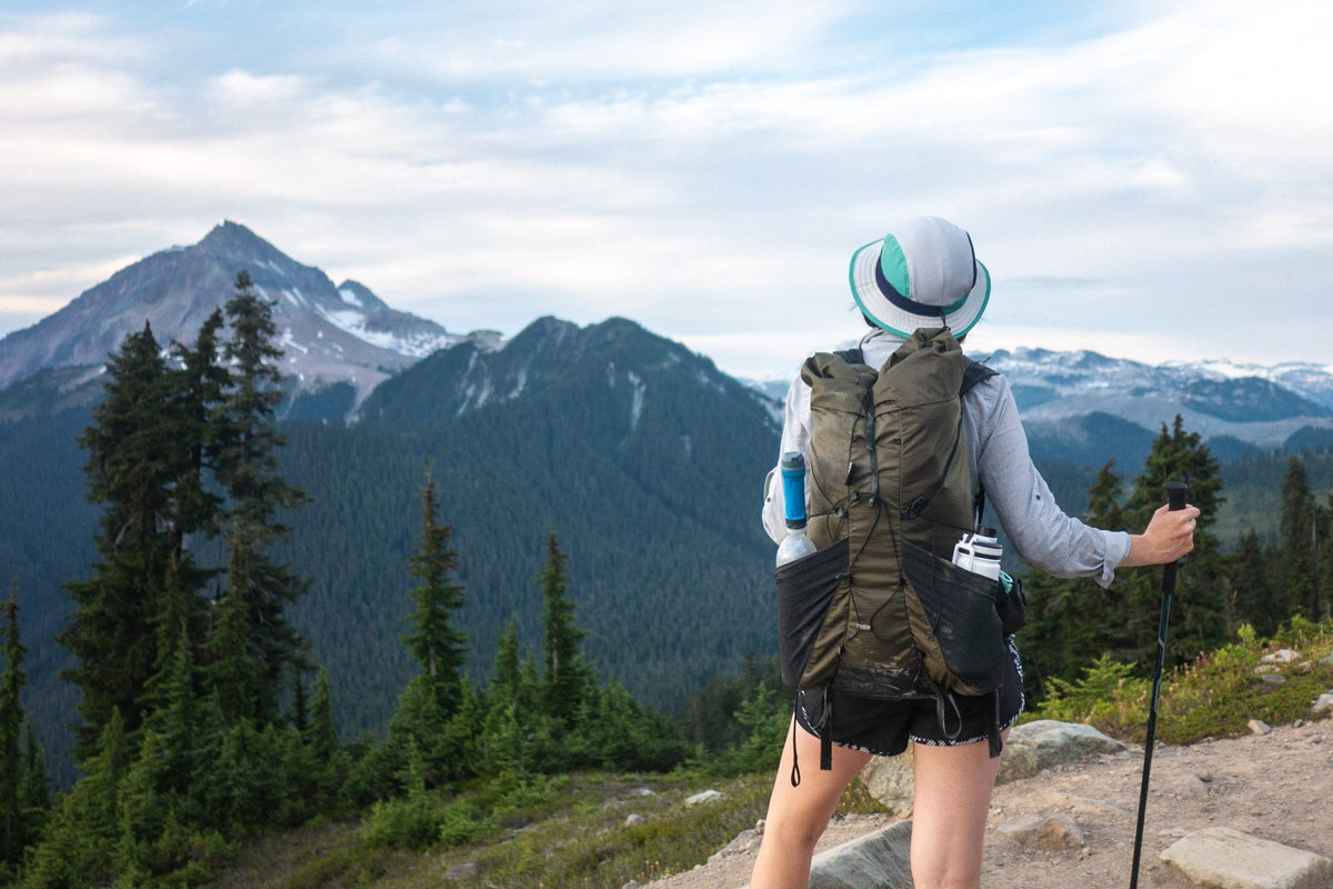 Hiker with a Flight 30 backpack and trekking pole looking at a glaciated peak above evergreen forest