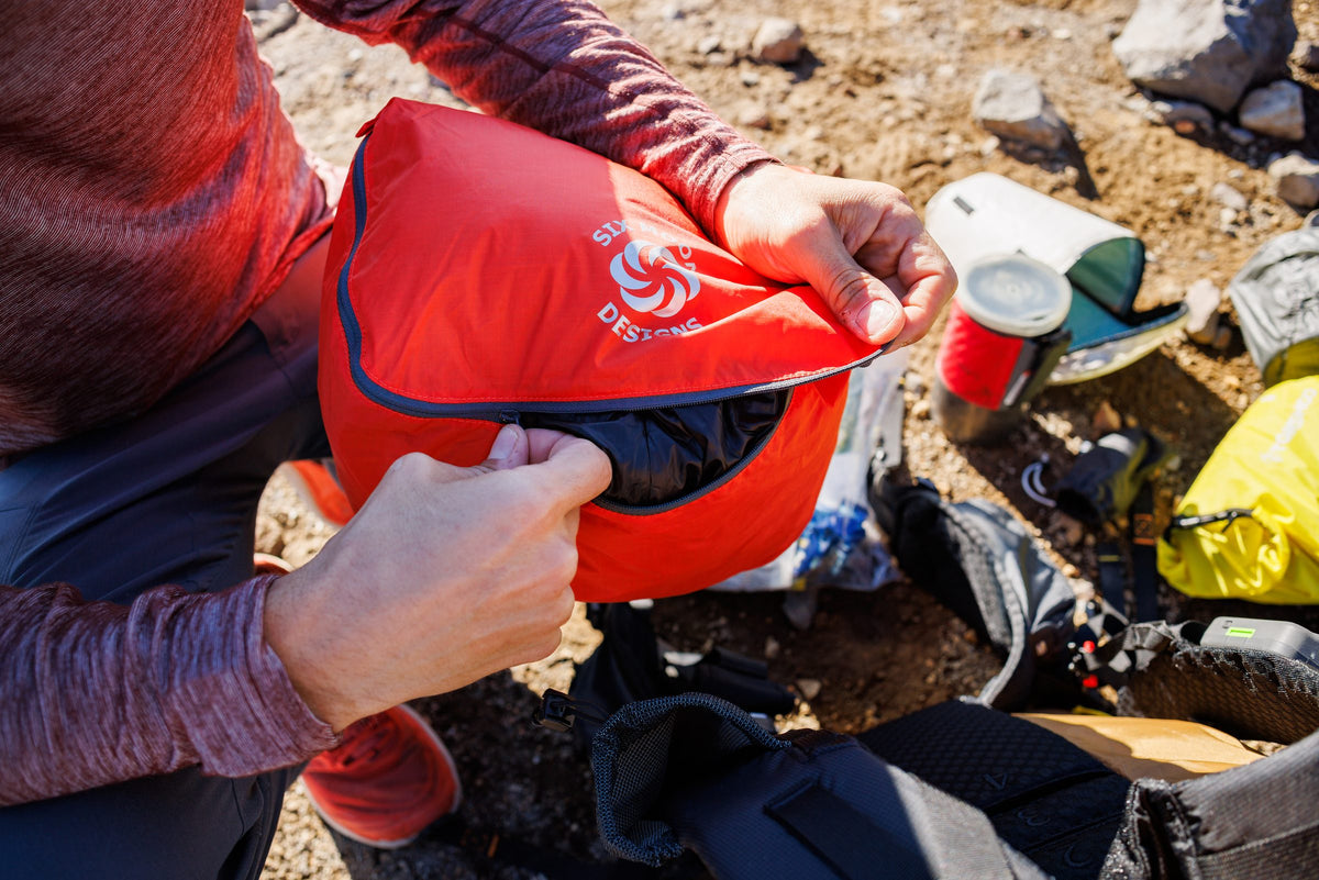 Hiker zipping an orange packing pod beside a backpack at a rocky campsite
