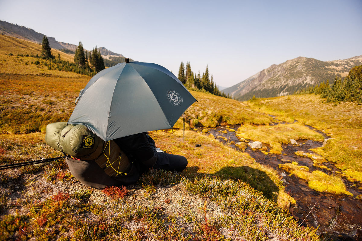 Hiker kneeling by an alpine creek in the shade of a dark teal Rain Walker SUL umbrella near Mount Rainier