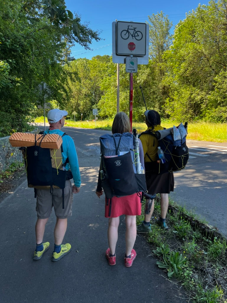 3 hikers wearing backpacks look at road sign for the Corvallis 2 Coast Trail