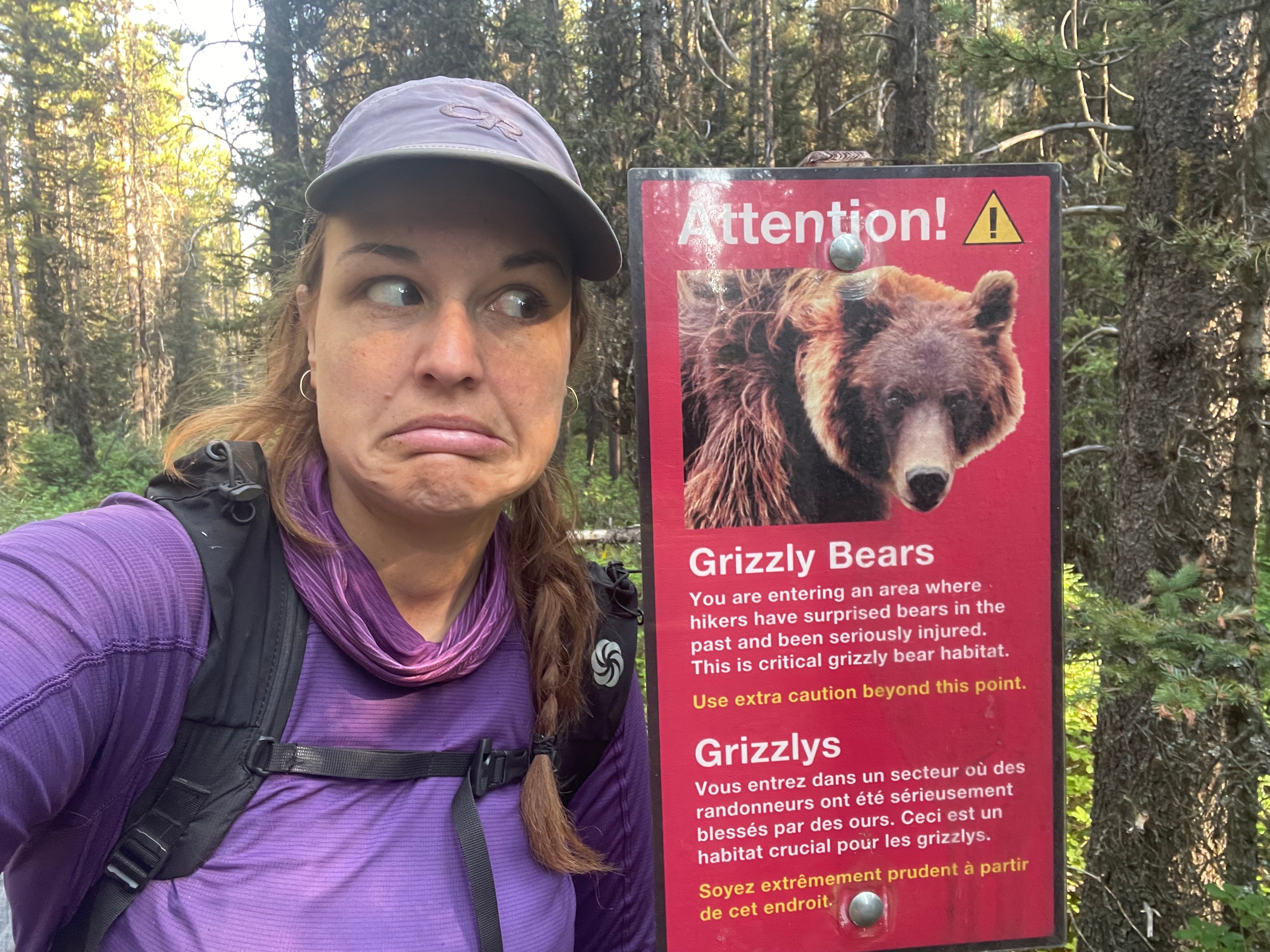 A hiker with a big frown on their face standing next to a sign warning about grizzly bears