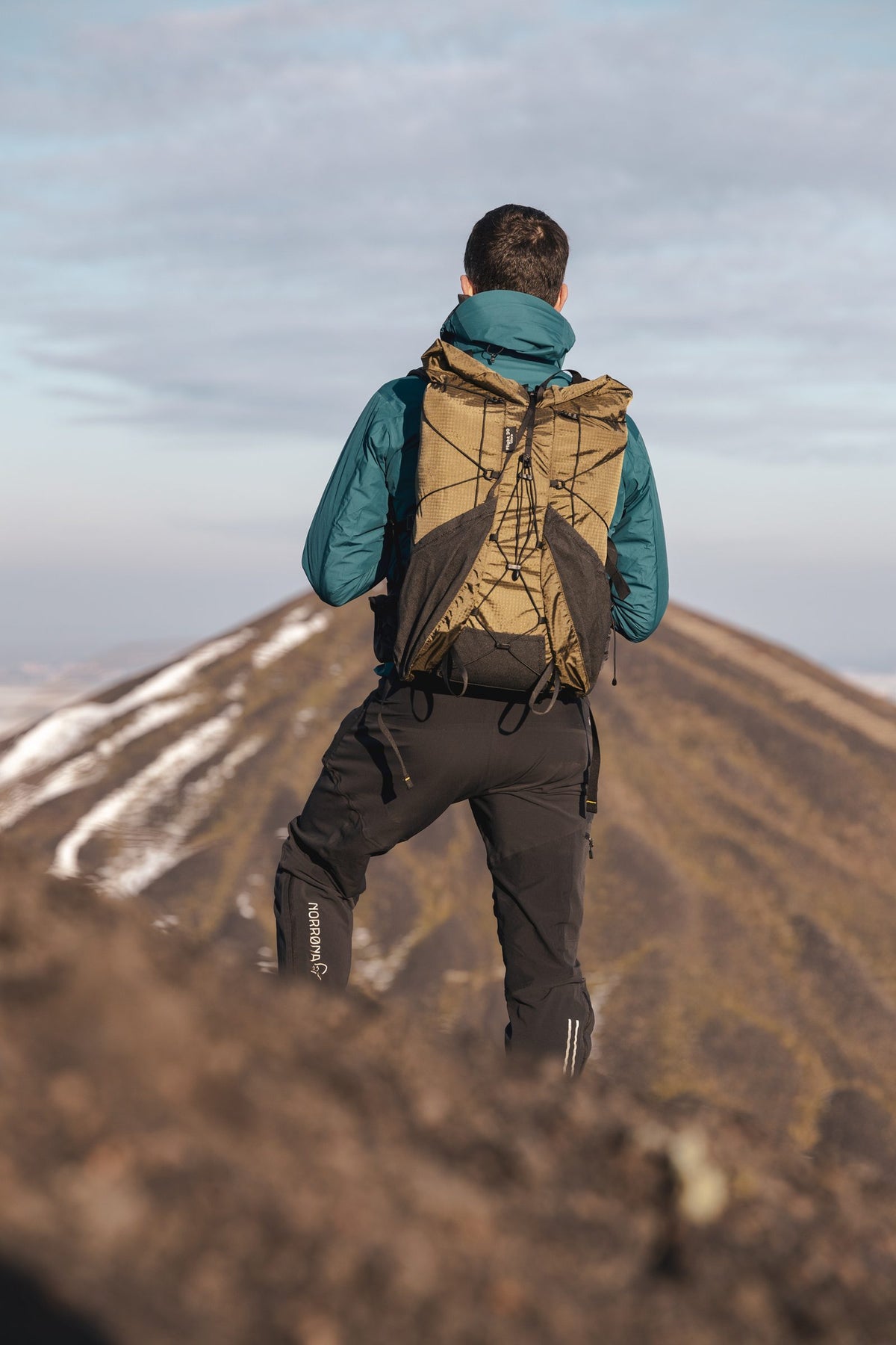 Hiker in a teal jacket wearing a coyote Flight 30 backpack climbing a volcanic ridge