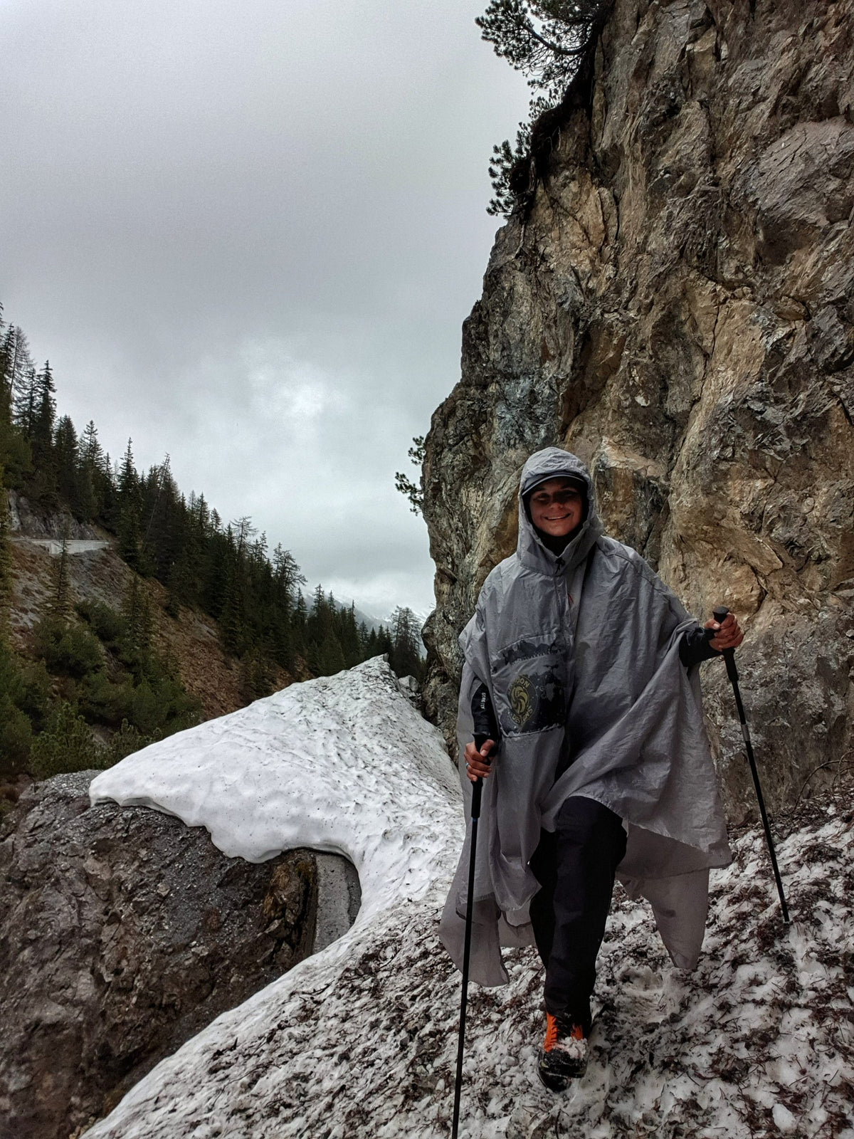 Hiker wearing the gray Gatewood Cape as a rain poncho on a snowy cliffside trail