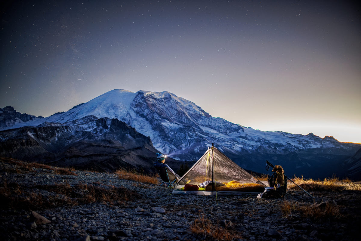 Two backpackers beside a lit Haven tent at twilight with Mount Rainier behind