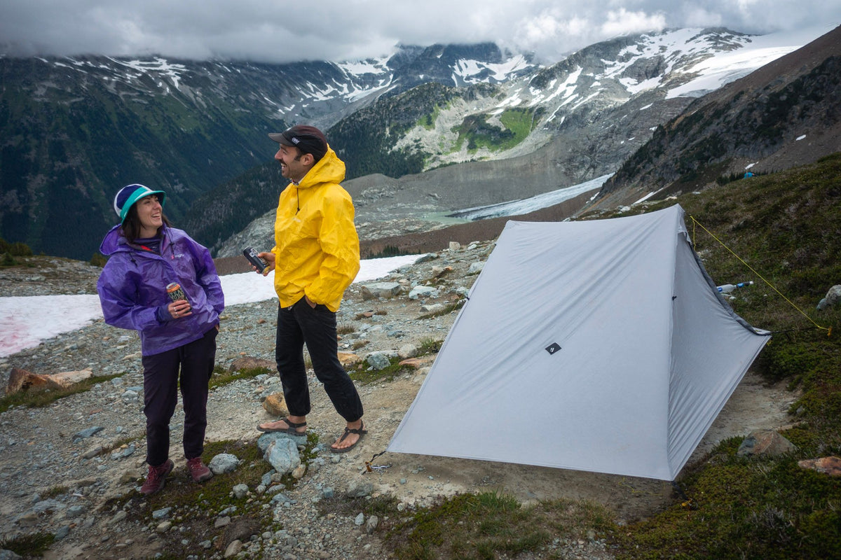 Two hikers chatting beside a gray Owyhee tarp on a rocky alpine slope above glaciers