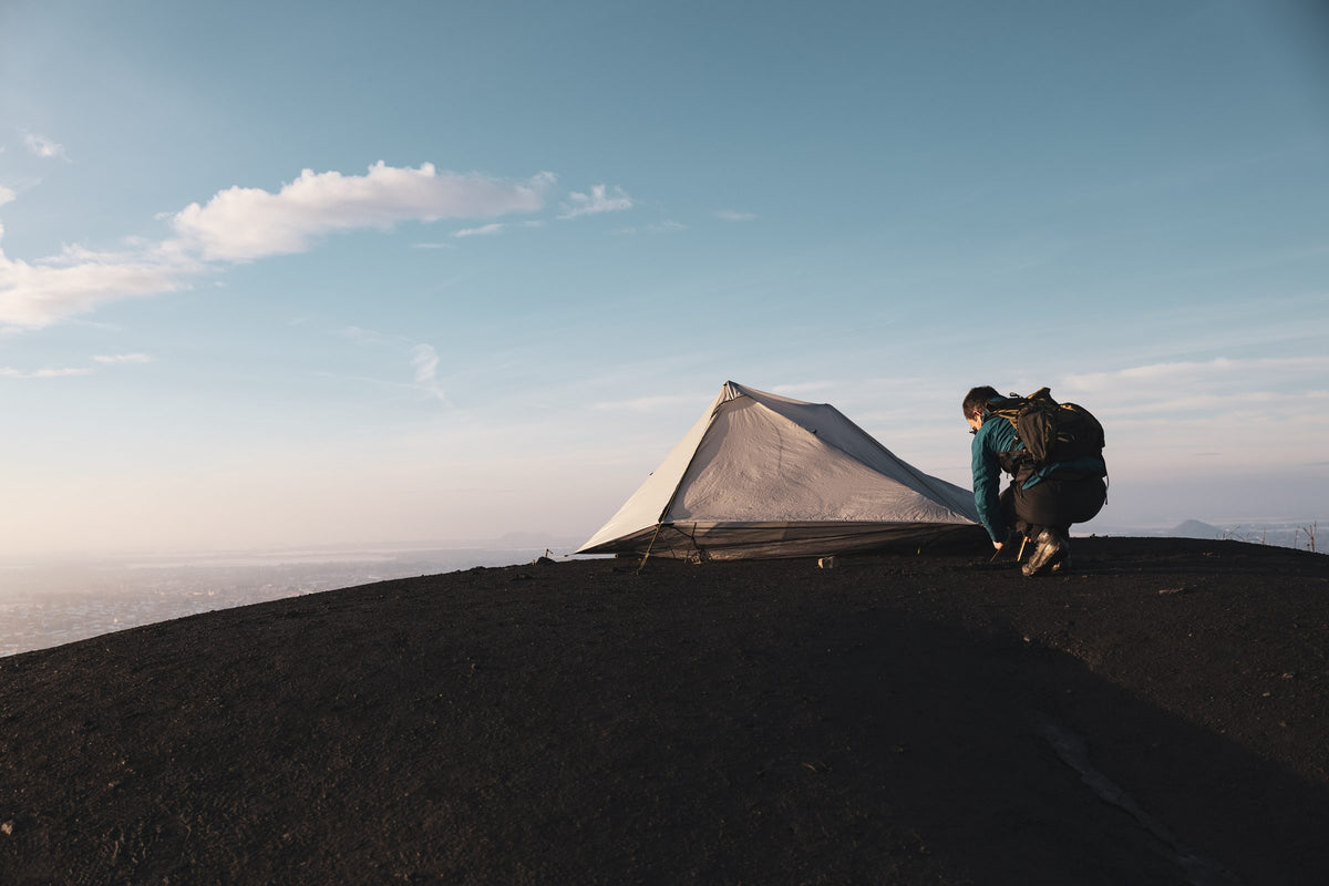 Hiker staking out a gray Owyhee tarp on a dark volcanic ridge at sunrise
