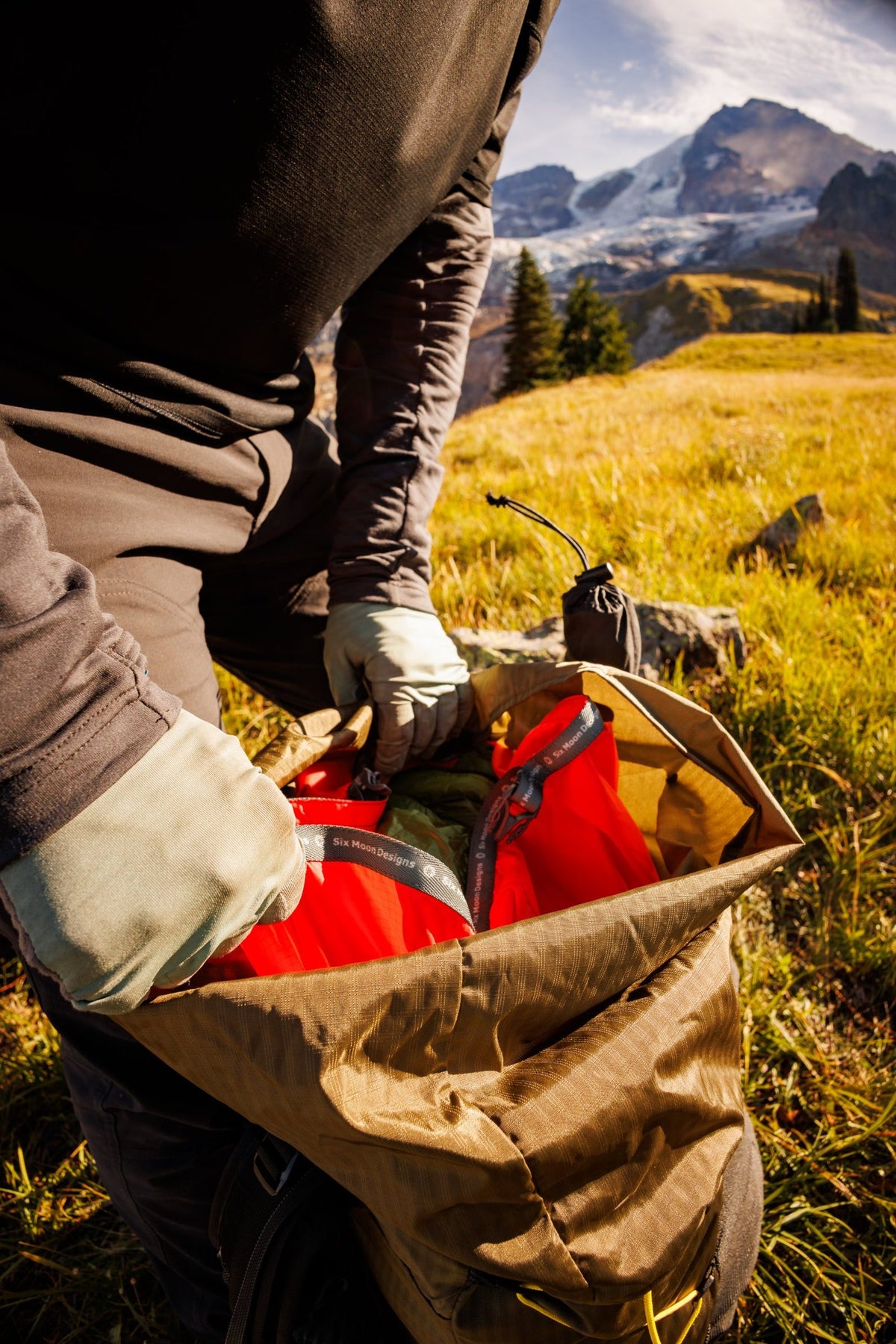 Hiker loading a red Six Moon Designs Pack Liner inside a roll-top backpack in an alpine meadow below Mount Rainier