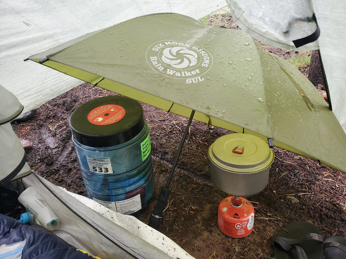 Olive Rain Walker SUL umbrella sheltering a camp kitchen with a bear canister, pot and stove in the rain
