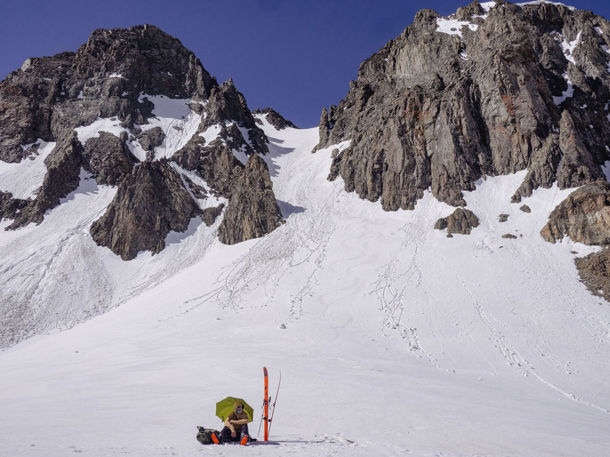 Backcountry skier sheltering under an olive Rain Walker SUL umbrella in a snowy couloir