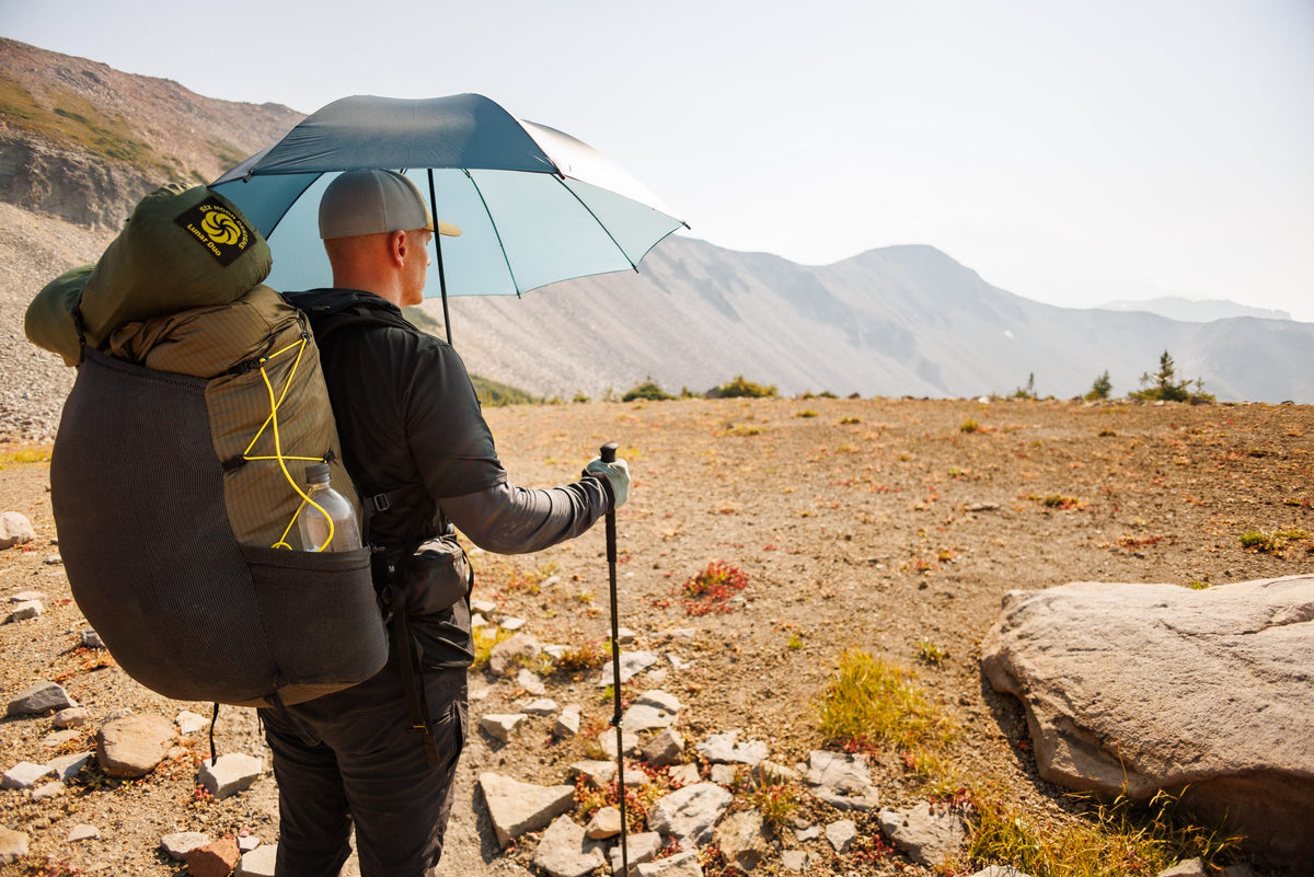 Hiker with a backpack holding a dark teal Rain Walker SUL umbrella on a rocky alpine ridge