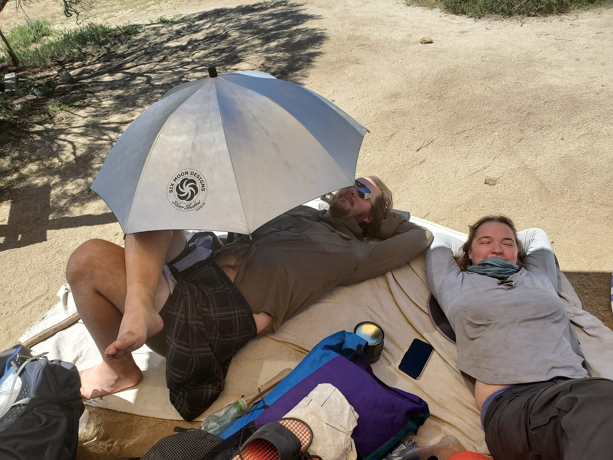Two hikers resting on sleeping pads in the shade of a Silver Shadow Carbon umbrella