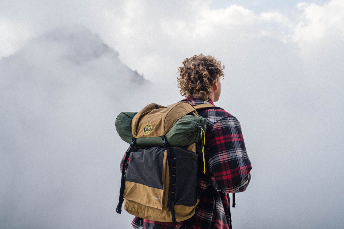 Hiker in flannel wearing a coyote brown Wy'east daypack with a sleeping pad strapped on, in misty clouds