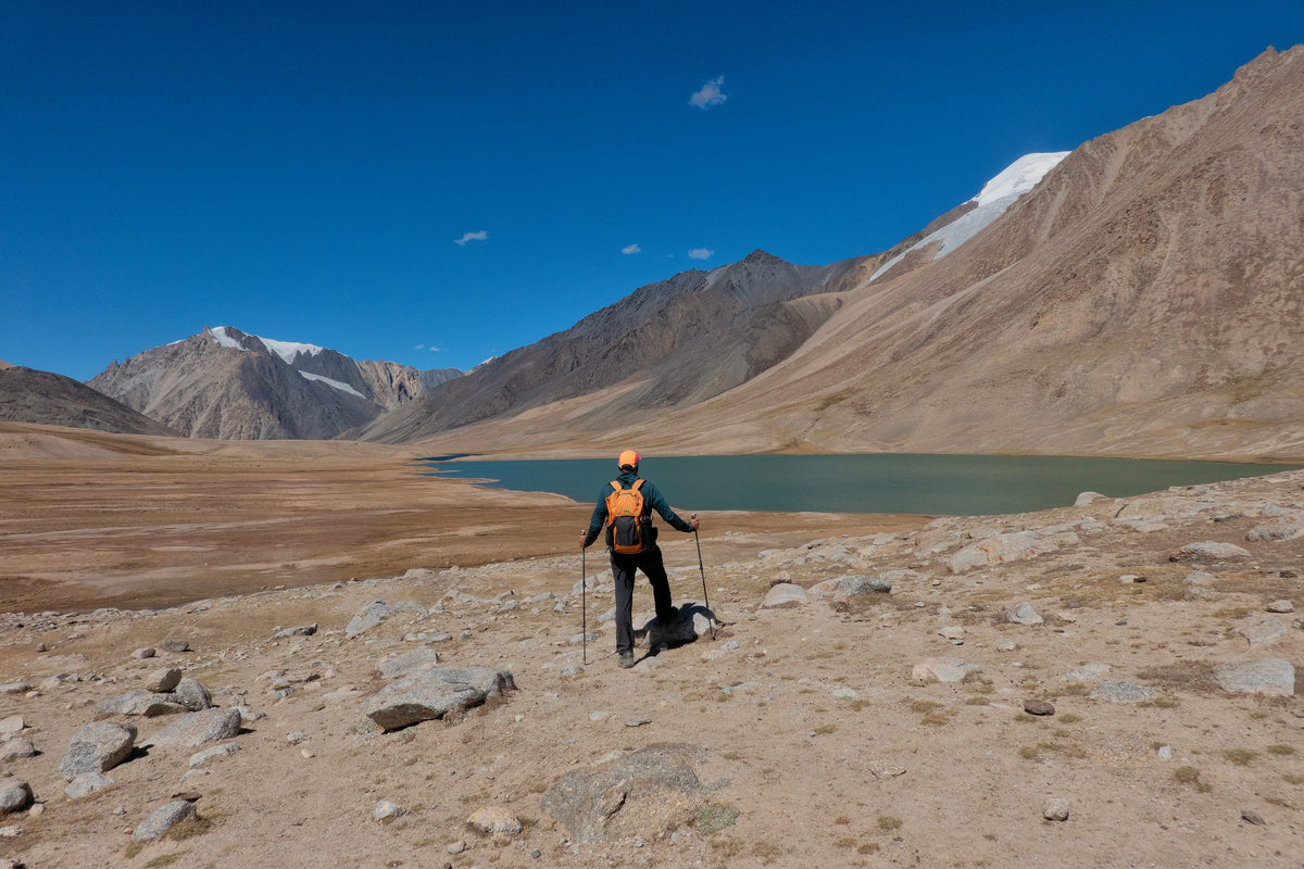 Hiker with an orange Wy'east daypack and trekking poles beside a high lake near Shimshal Pass, Pakistan