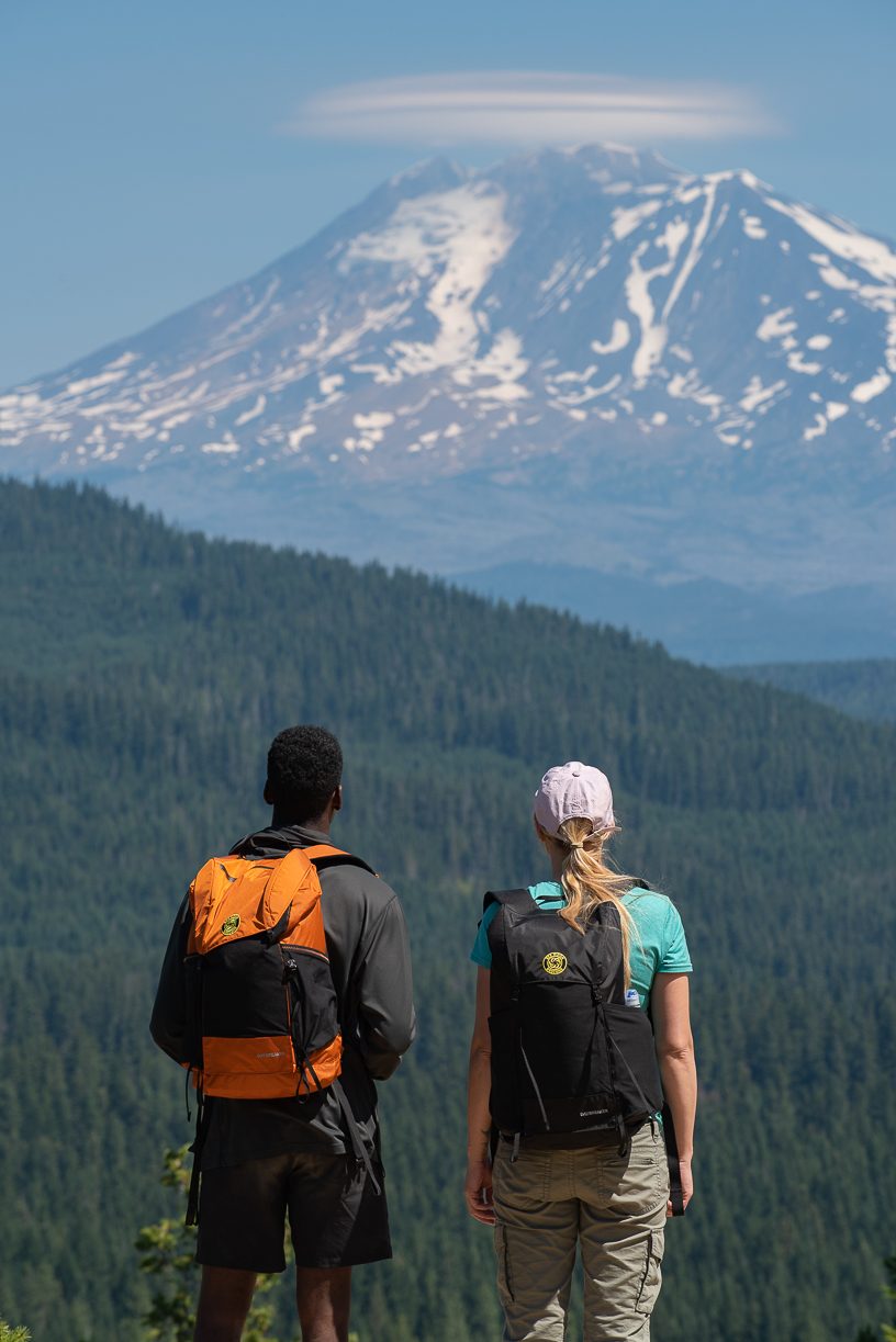Two hikers with orange and black Wy'east daypacks looking out at a snow-capped volcano